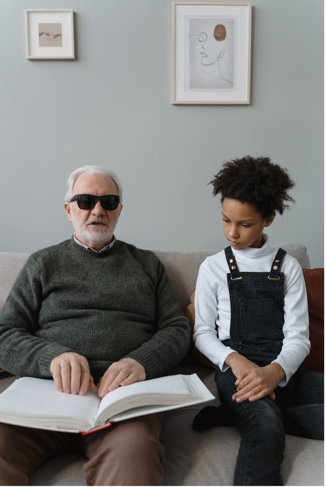 A man with a white beard and dark glasses is sitting on a sofa. Next to him is a young girl aged around 10. He is holding a Braille book and is reading to her.